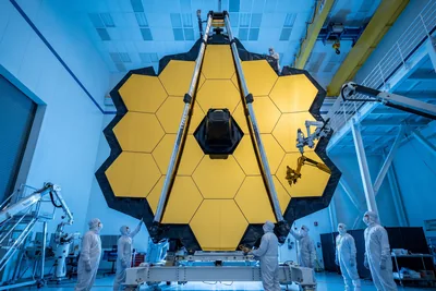 Illustration éditoriale d’un grand télescope spatial à miroir hexagonal doré assemblé dans une salle blanche, entouré d’ingénieurs en combinaison